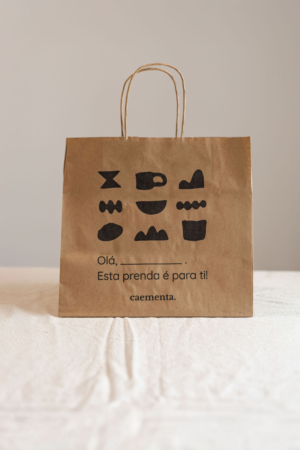Saco de papel castanho com ilustrações pretas e texto em português colocado sobre tecido branco.