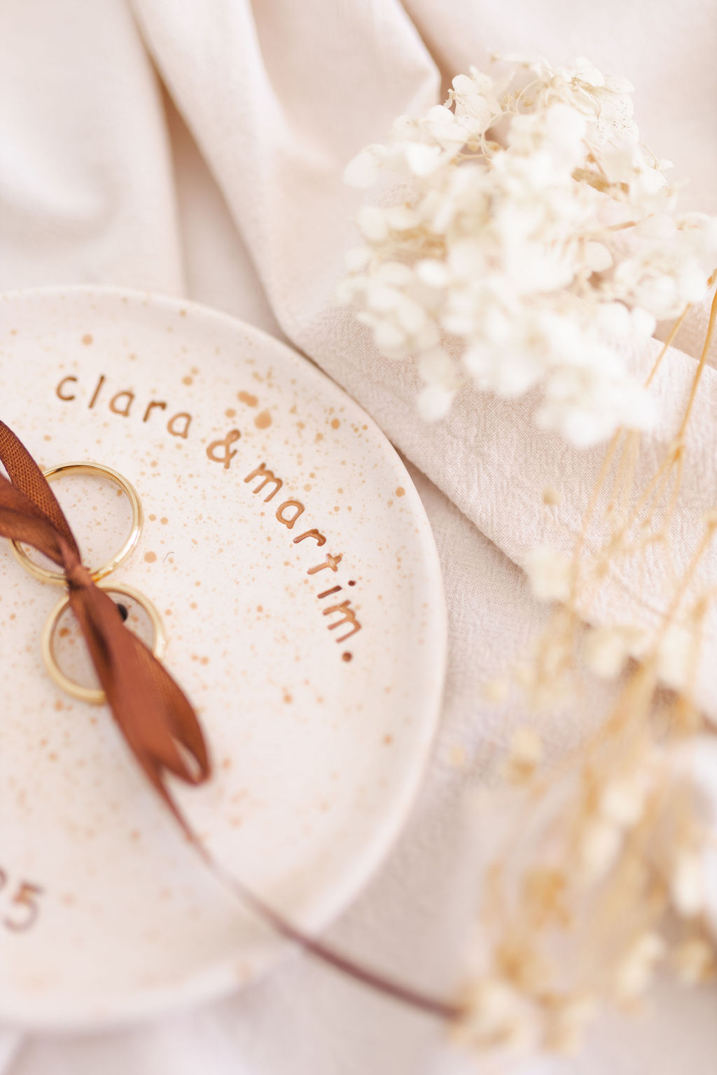 Prato cerâmico branco com nomes gravados e anéis dourados com fita castanha sobre tecido bege e flores secas.