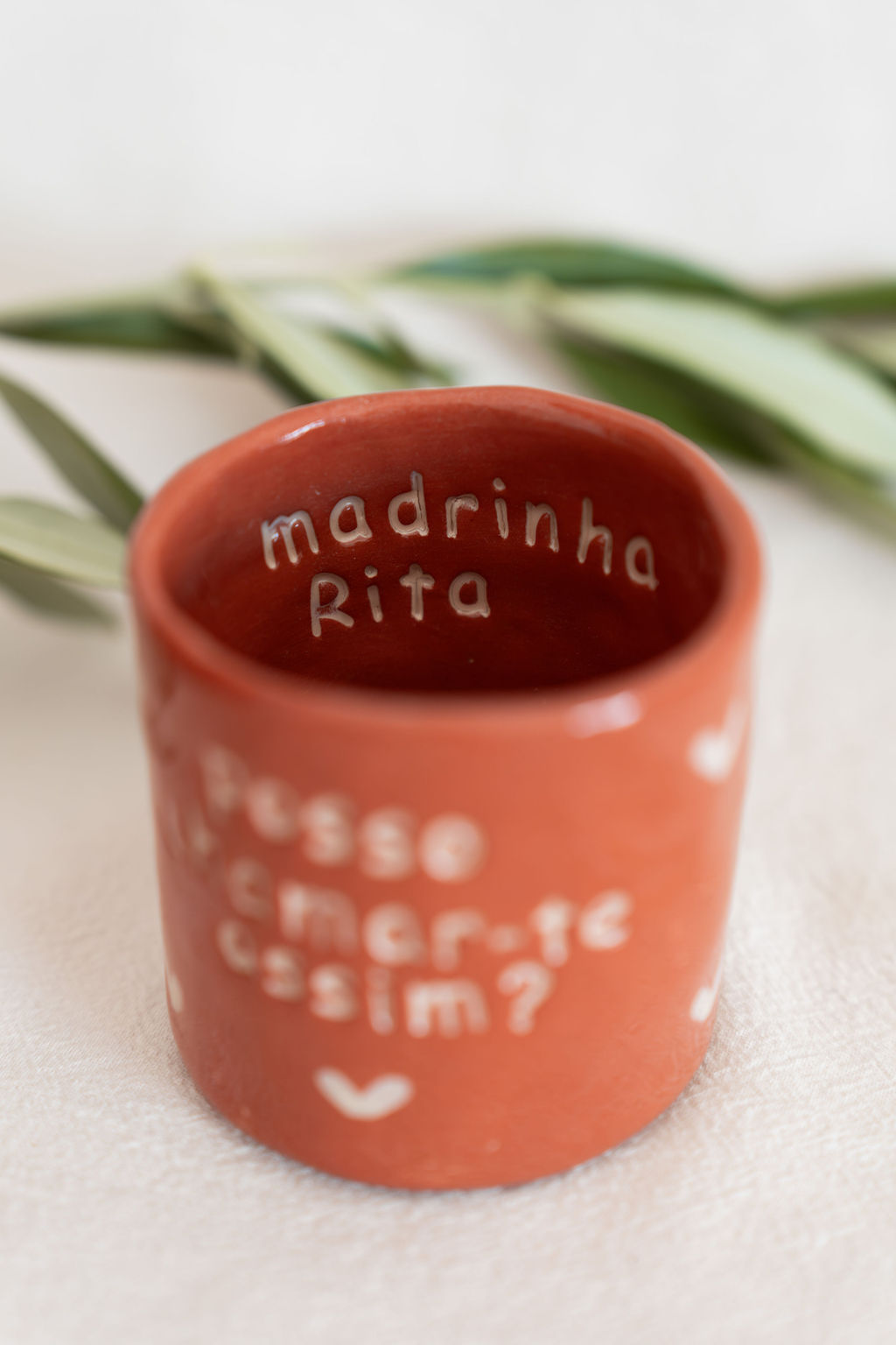 Copo de cerâmica castanho com texto branco pintado e folhas verdes ao fundo