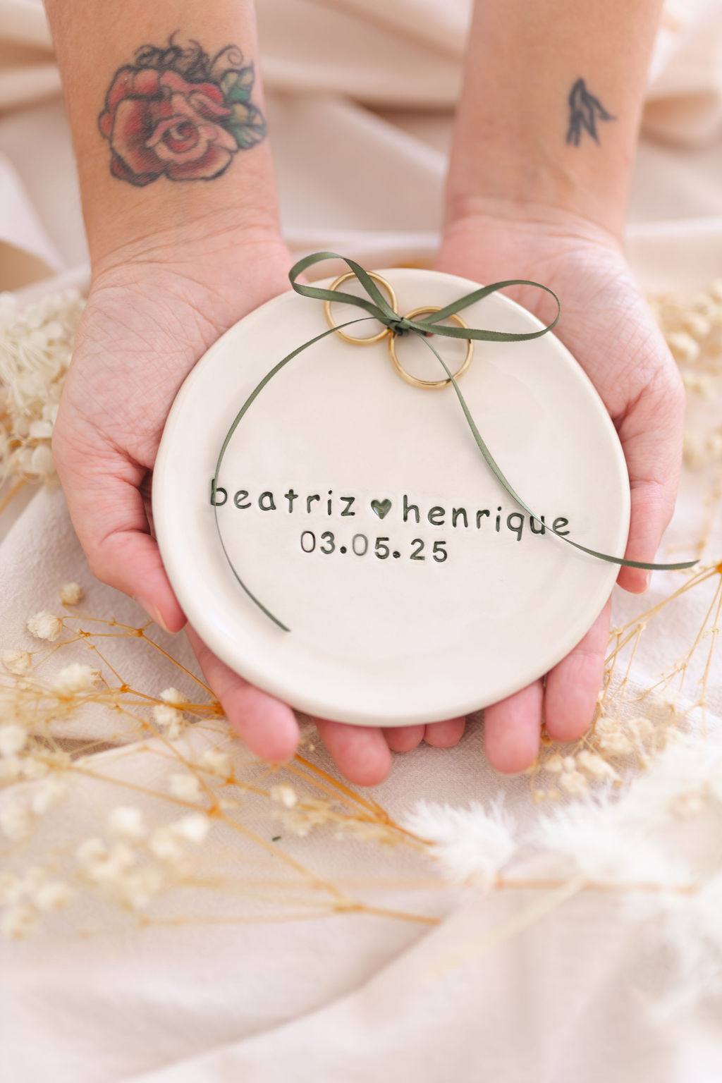 Prato cerâmico branco com nomes e data, alianças douradas com fita verde, mãos tatuadas seguram o prato