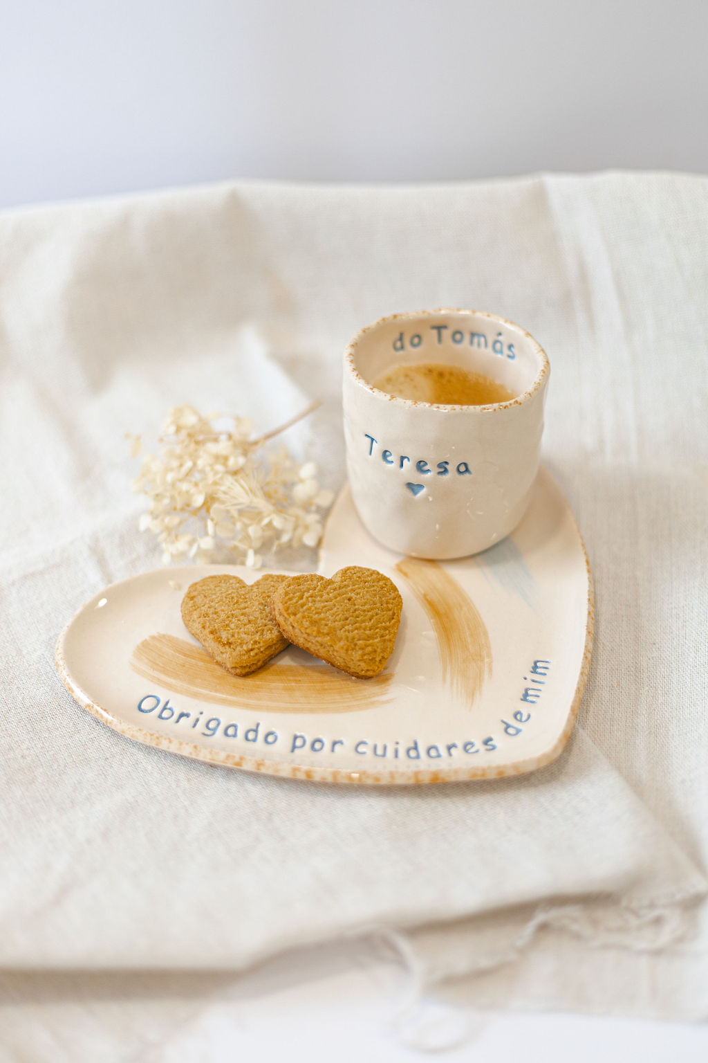 Conjunto cerâmico com chávena e prato em forma de coração em tons creme, com texto personalizado em azul e biscoitos em forma de coração.