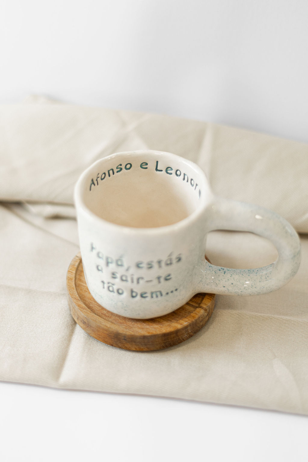 Caneca de cerâmica creme com texto azul, base de madeira e pano bege