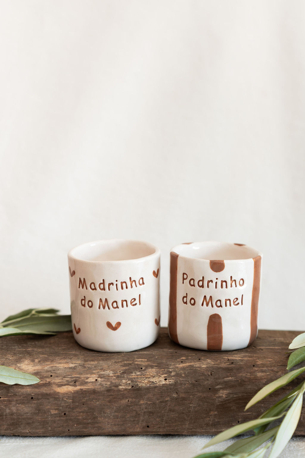 Dois vasos de cerâmica branca com texto em castanho Madrinha do Manel e Padrinho do Manel sobre madeira com folhas verdes