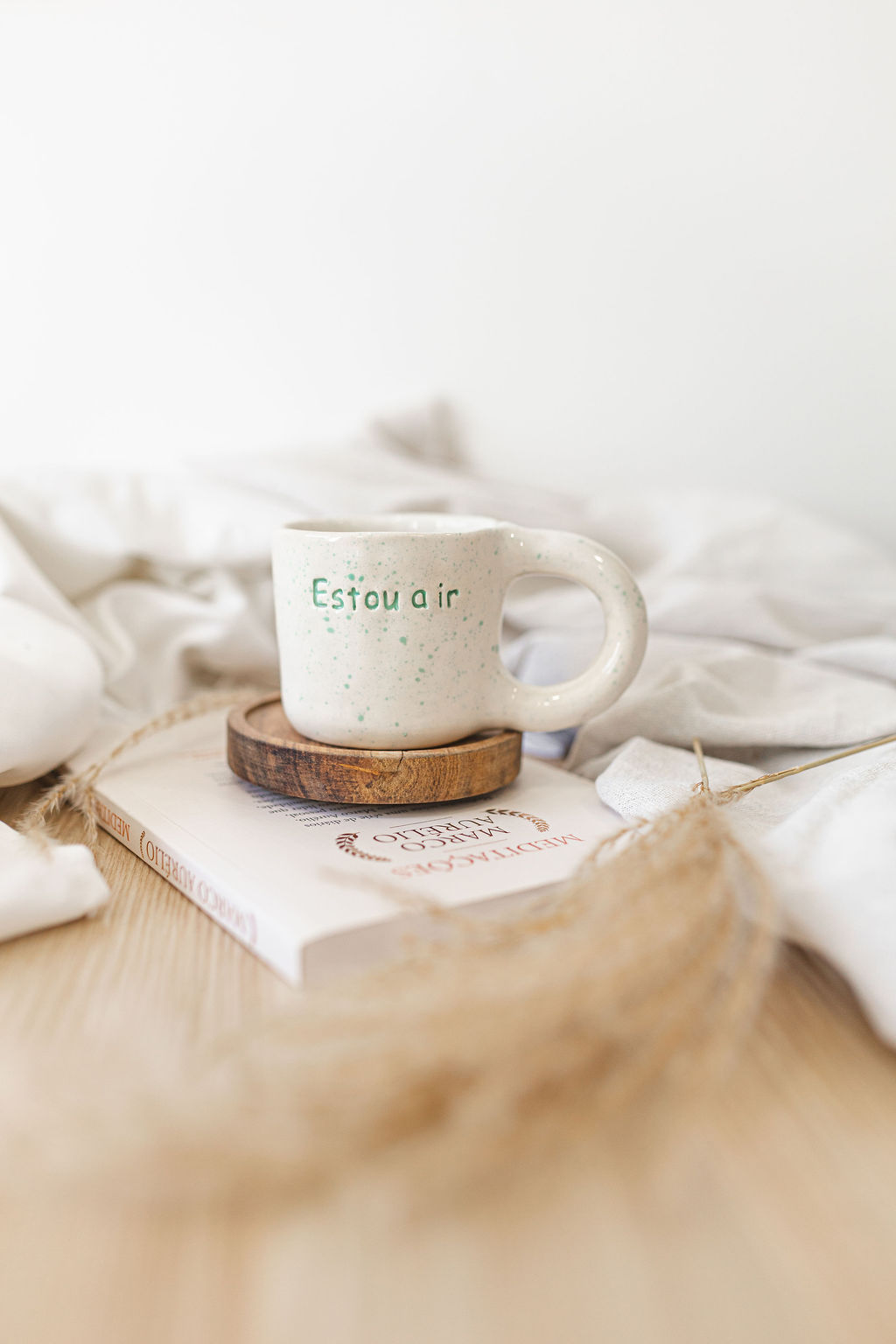 Caneca de cerâmica branca com texto verde sobre base de madeira e livro com tecido ao fundo
