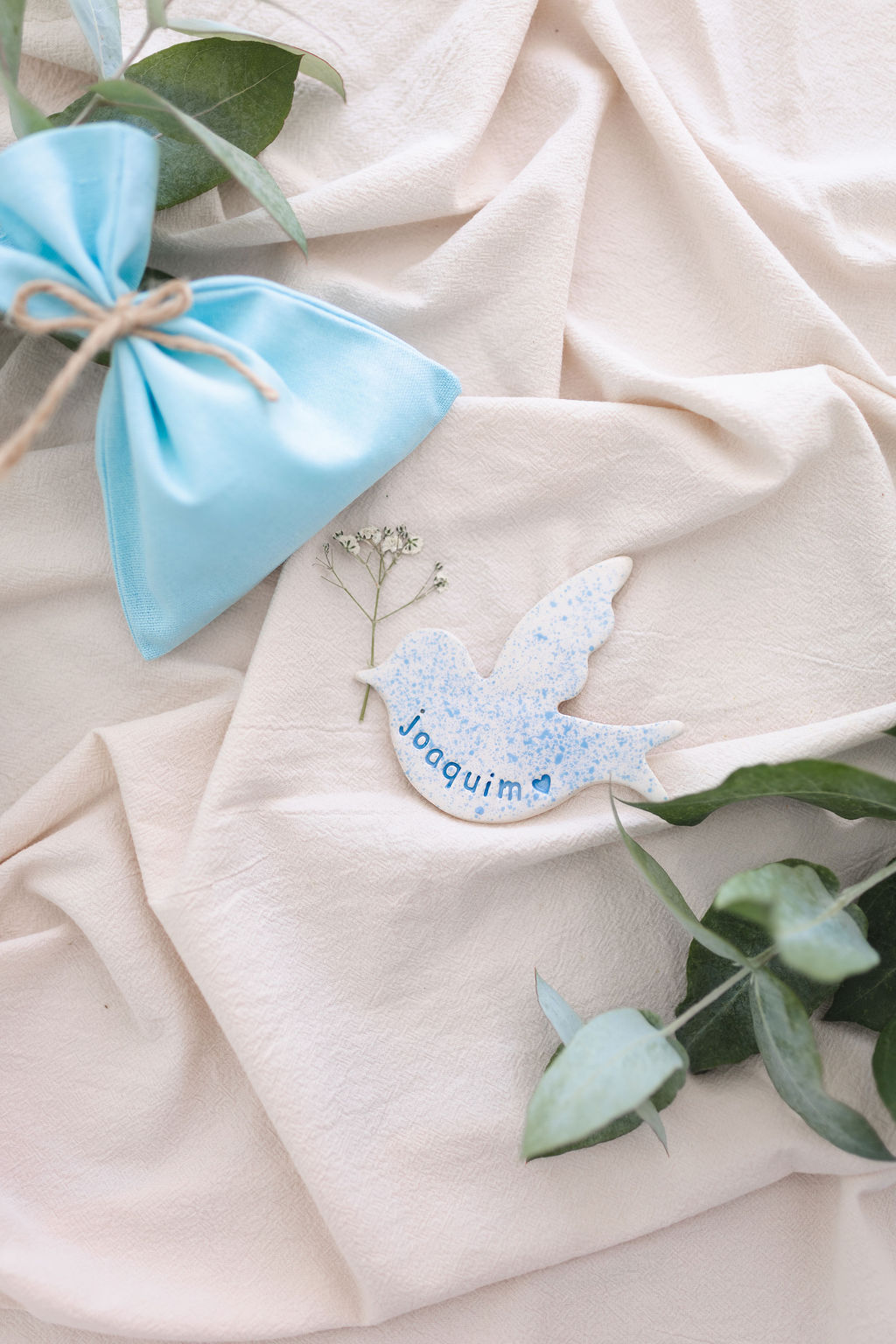 Pássaro decorativo em cerâmica azul sobre tecido creme com folhas verdes e saco azul amarrado.