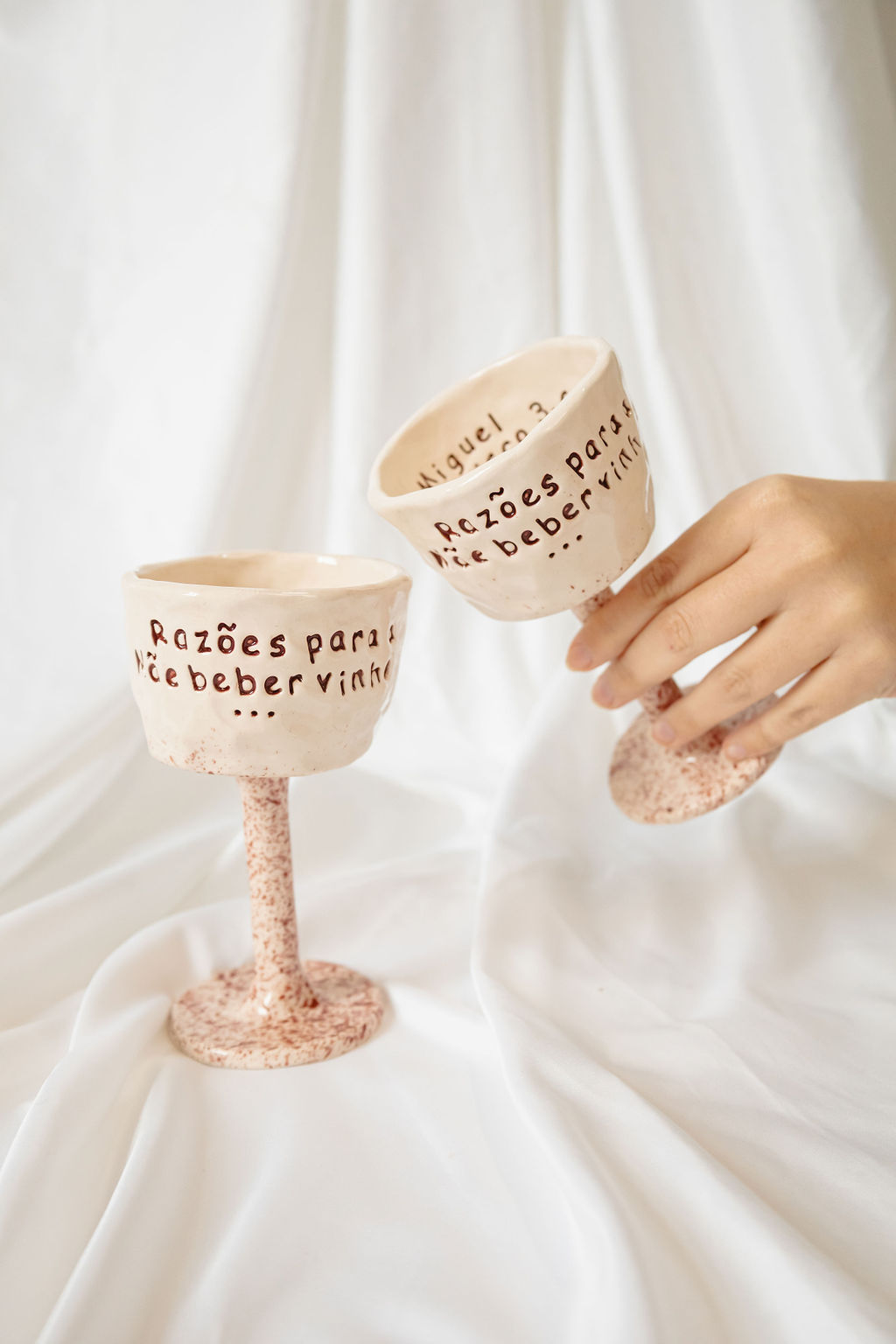 Duas taças de vinho em cerâmica com inscrição em português e acabamento craquelado, uma sendo segurada por uma mão
