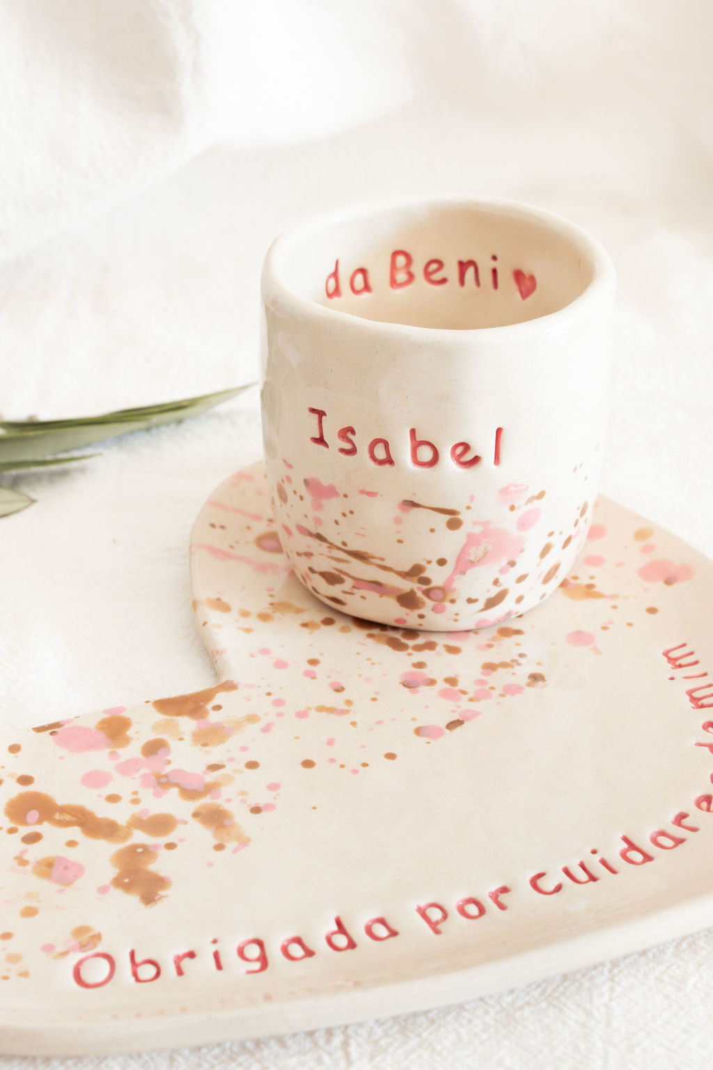 Conjunto cerâmico branco com splashes cor de rosa e castanho e texto em vermelho, composto por copo e prato.