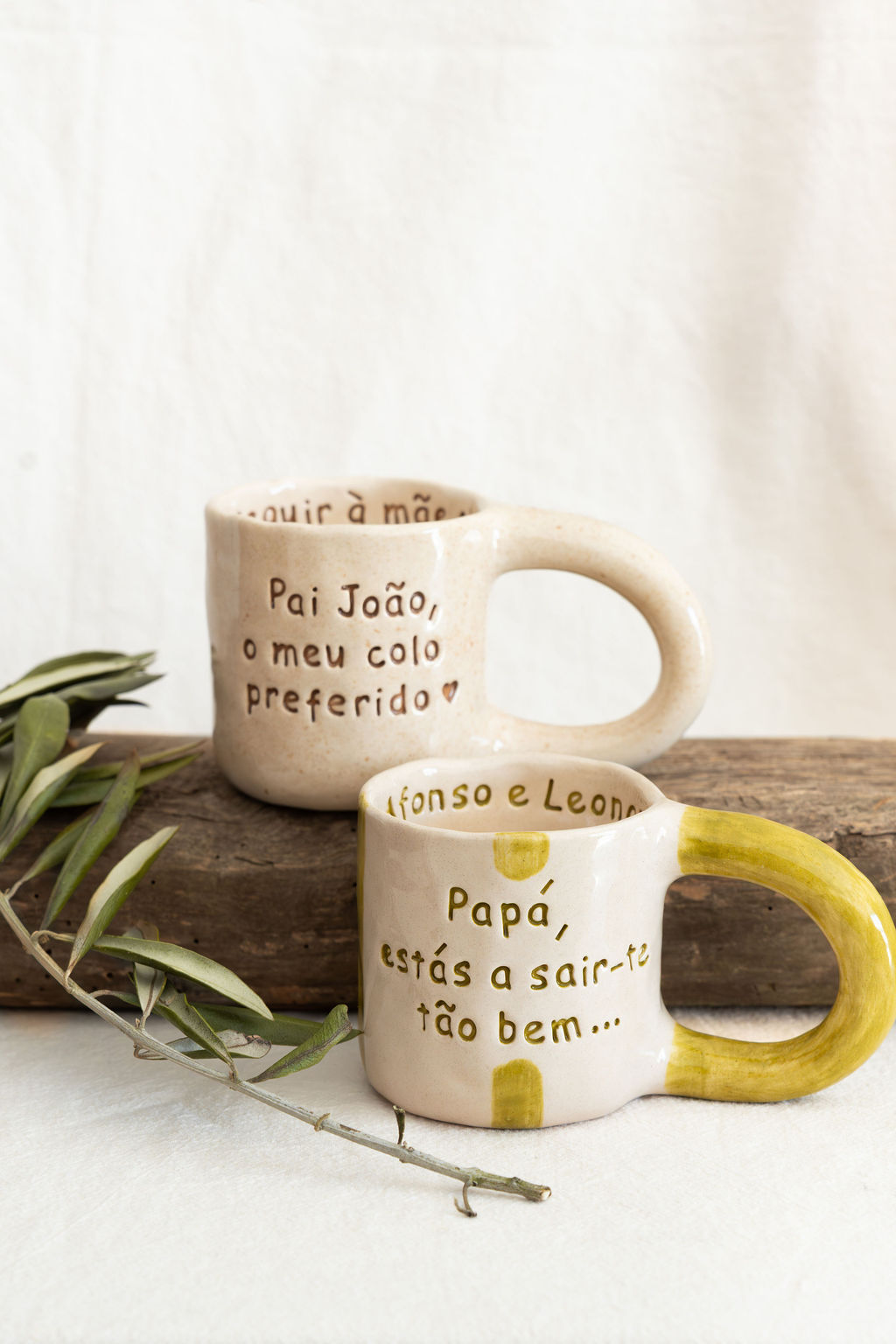 Duas canecas de cerâmica bege com texto em português, uma com alça amarela, em fundo bege e madeira com ramo verde