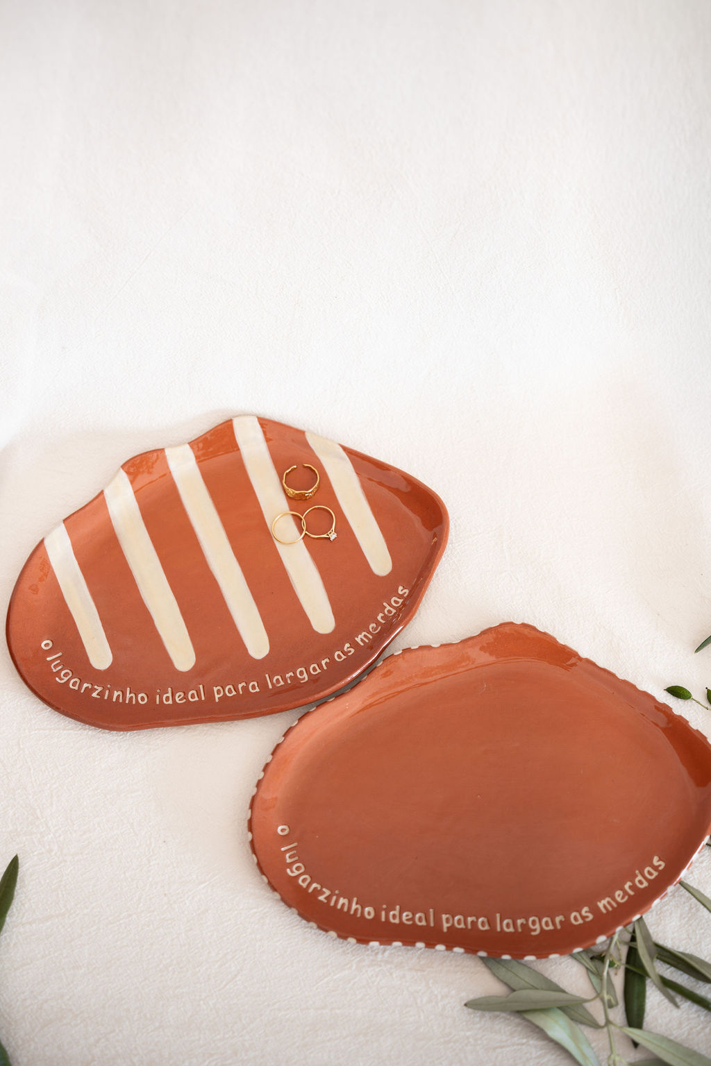 Duas pequenas bandejas de cerâmica terracota, uma com riscas brancas e anéis dourados, com escrita em branco.
