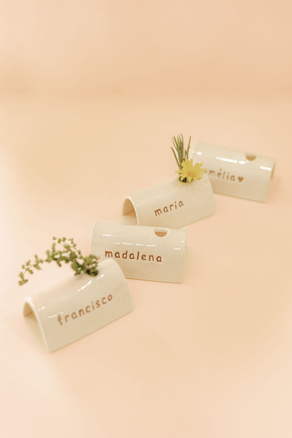 Vasos cerâmicos brancos com nomes escritos e pequenas flores em fundo cor de pêssego
