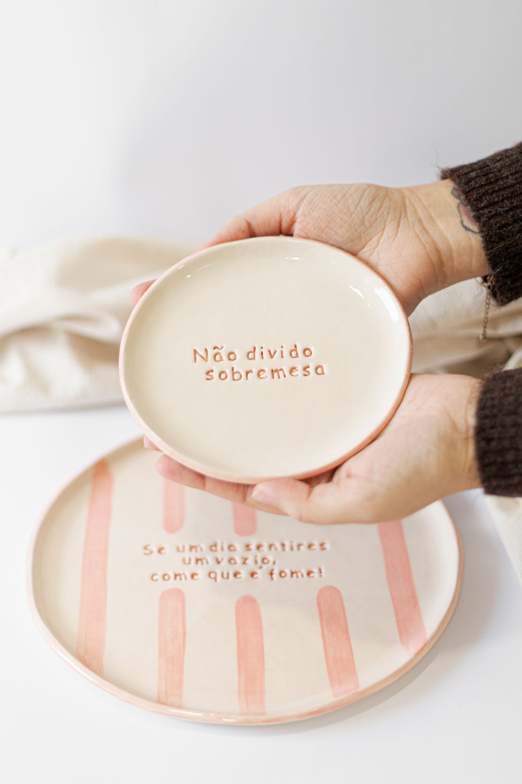 Dois pratos de cerâmica com texto em português, prato pequeno nas mãos com a frase Não divido sobremesa e prato maior ao fundo com riscas castanhas e outra mensagem.