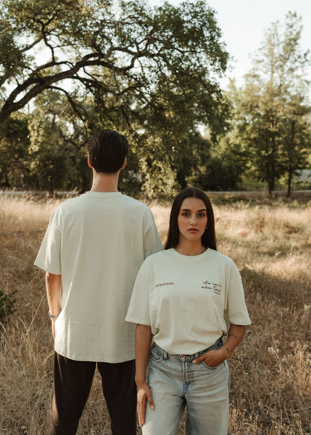 Dois modelos com t-shirts brancas e calças ao ar livre junto a uma árvore grande