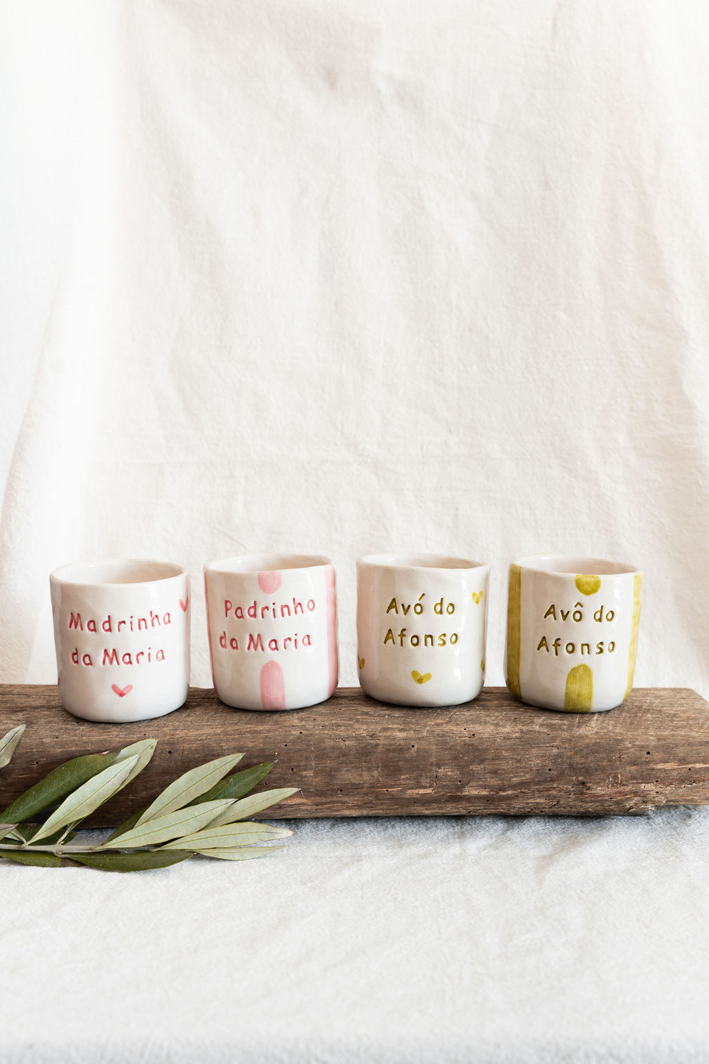 Quatro copos de cerâmica branca com textos em rosa e dourado sobre um suporte de madeira e fundo branco.