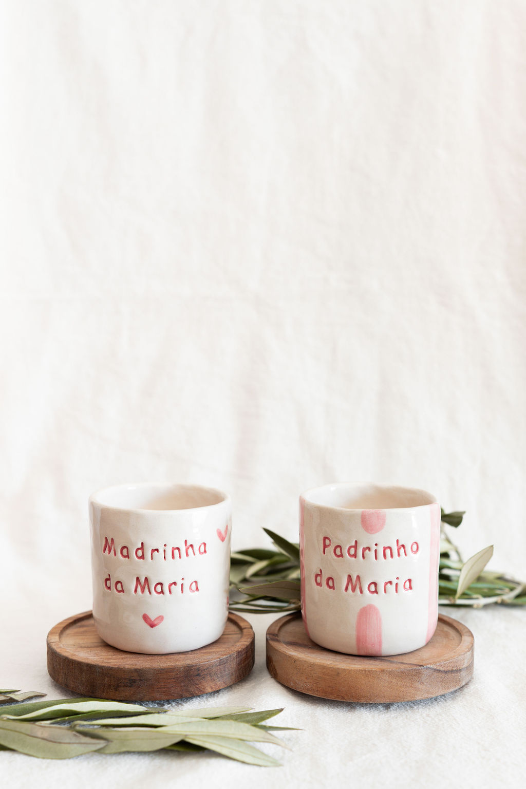 Duas canecas de cerâmica branca com texto vermelho em cima de bases de madeira com folhas verdes ao redor.