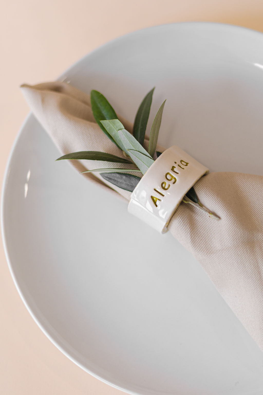 Guardanapo bege com anel cerâmico branco Allegria e raminho verde num prato branco.