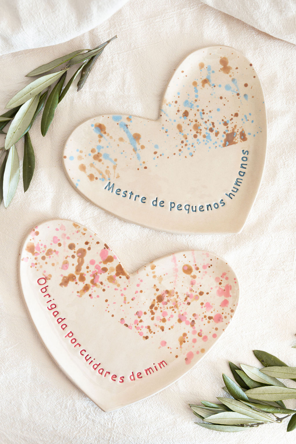 Dois pratos em forma de coração com salpicos coloridos e frases em português, sobre tecido claro com folhas verdes