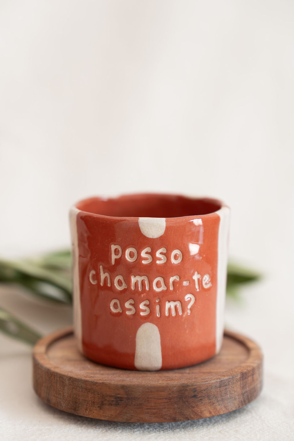 Caneca de cerâmica terracota com manchas brancas e texto em relevo, sobre suporte de madeira.