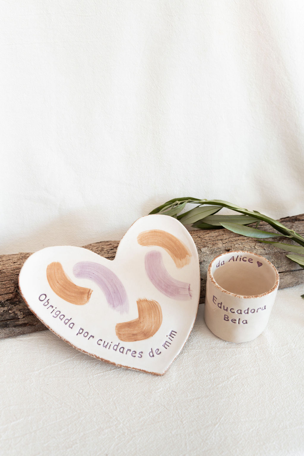Conjunto de prato em forma de coração e caneca de cerâmica com inscrições em português, decorados com pinceladas castanhas e lilás, cenário com tronco e folhas.