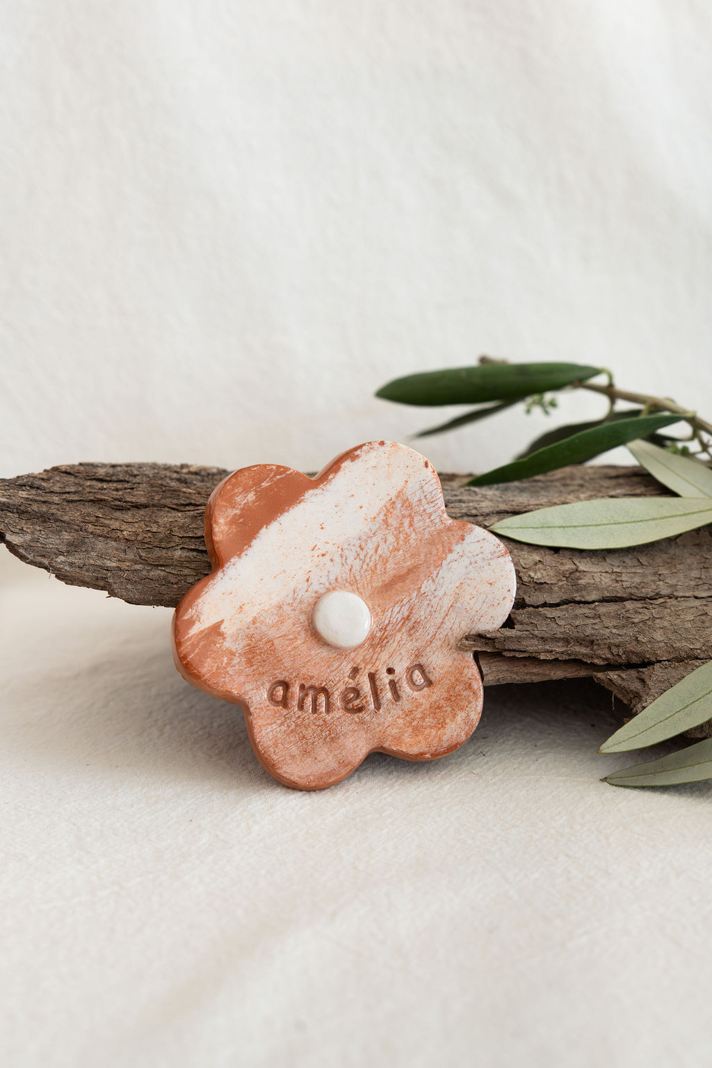 Broche de cerâmica em forma de flor com o nome amélia inscrito, posicionado sobre uma casca de árvore com folhas verdes ao lado.