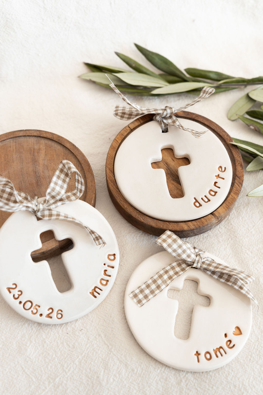 Adornos cerâmicos brancos com cruz recortada, nomes e laços xadrez sobre madeira e tecido.