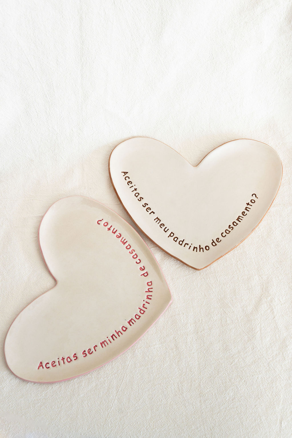 Dois pratos cerâmicos em forma de coração com texto dirigido a padrinho e madrinha de casamento sobre tecido branco.