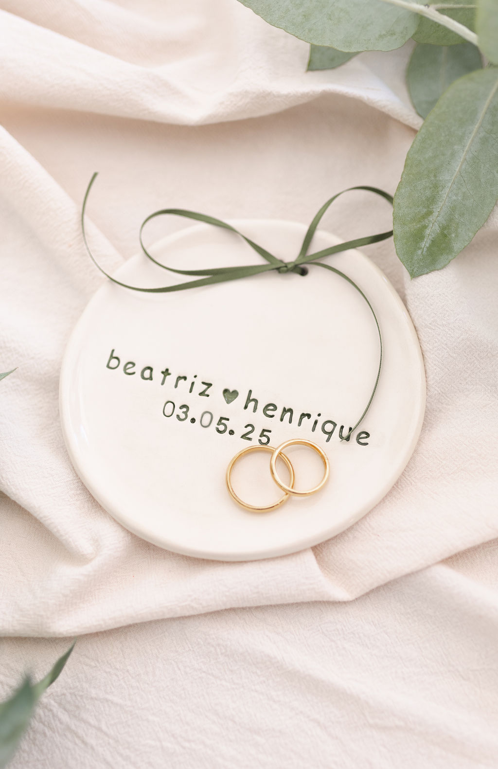 Prato branco de cerâmica com inscrições verdes, fitas e alianças douradas sobre tecido claro e folhas verdes