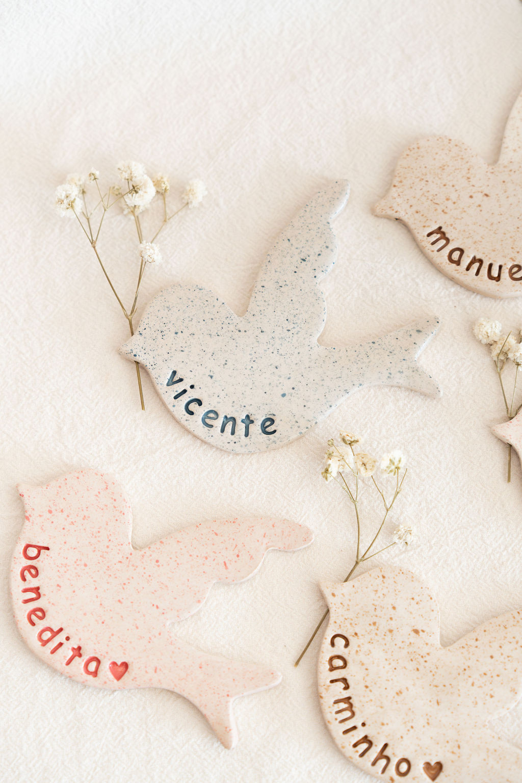 Pássaros decorativos de cerâmica com nomes pintados e flores secas sobre tecido branco.