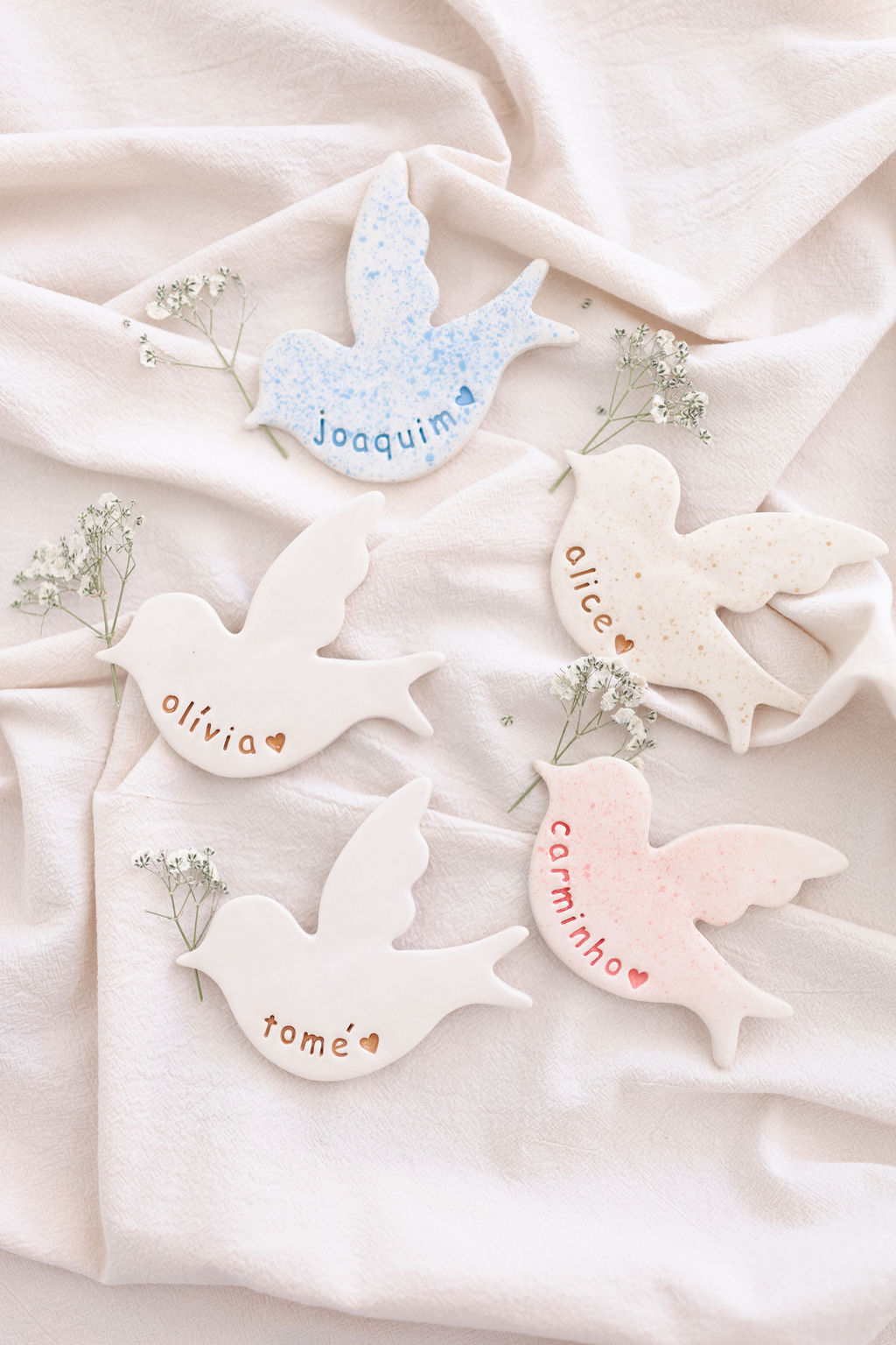 Cinco pássaros de cerâmica com nomes gravados e pequenos ramos de flores sobre tecido bege.