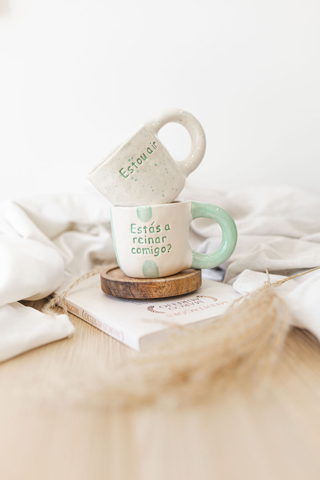 Duas chávenas de cerâmica branca e verde com texto em português, empilhadas sobre um prato de madeira e um livro