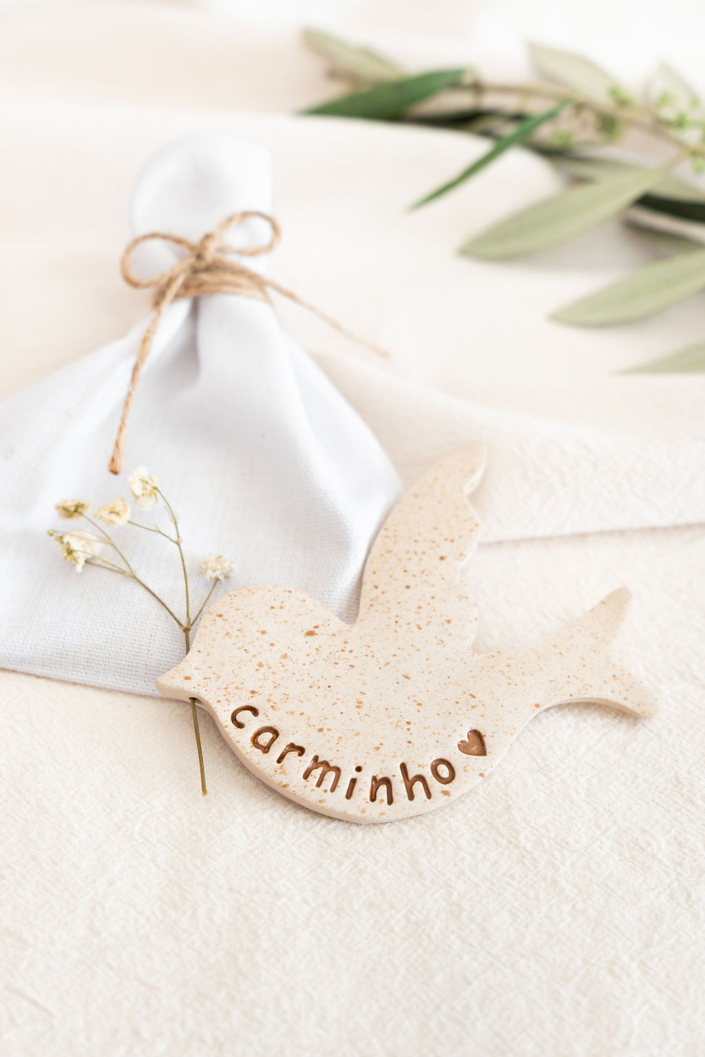 Porta-guardanapo em forma de pássaro de cerâmica bege com a palavra carminho e guardanapo branco com corda e flores secas