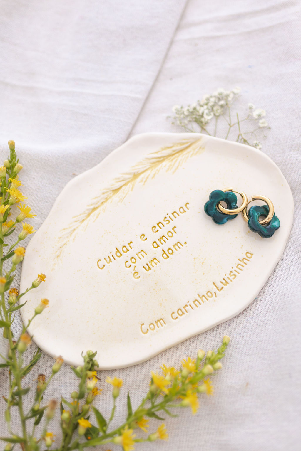 Brincos turquesa com detalhes dourados num prato branco decorado com texto em dourado, flores amarelas ao redor.