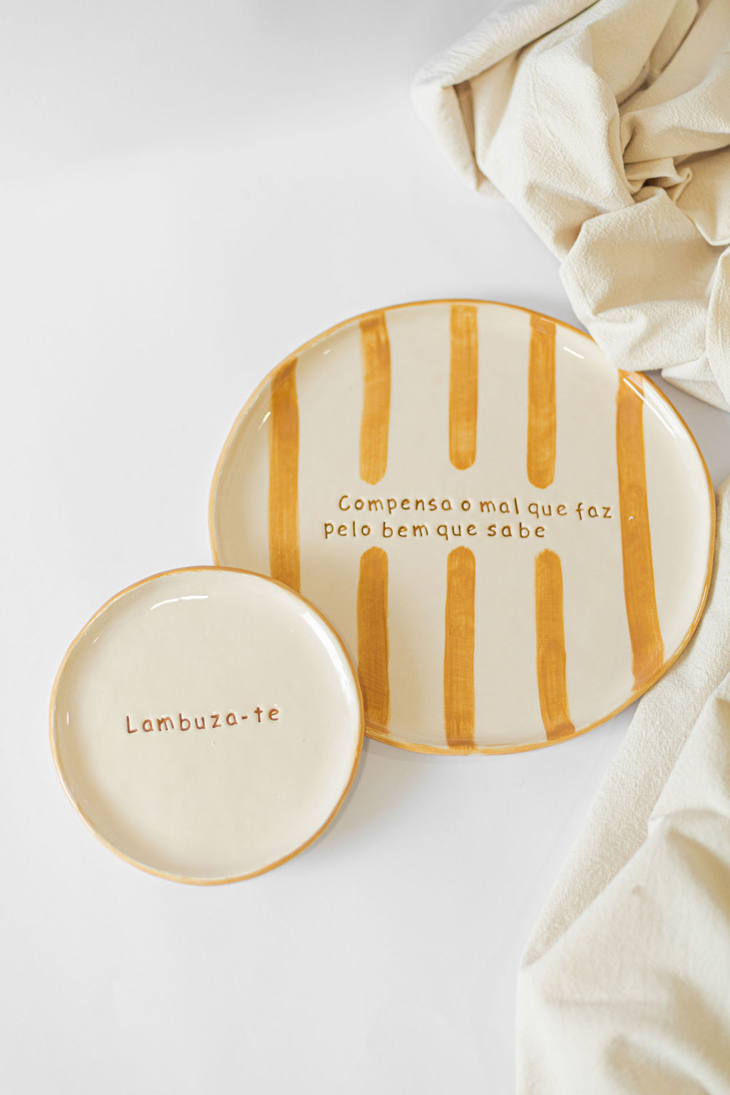 Dois pratos de cerâmica creme com texto em castanho e riscas castanhas, apoiados numa superfície branca junto a tecido bege.