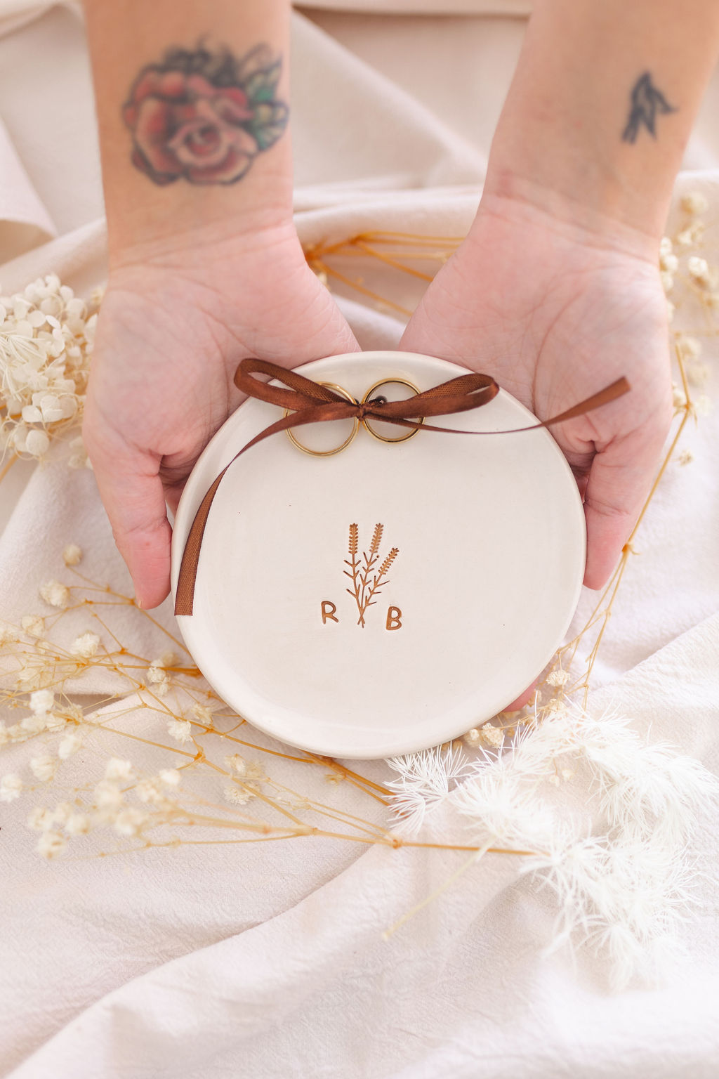 Prato cerâmico branco com alianças douradas amarradas com fita castanha, rodeado por flores secas e tecido claro.