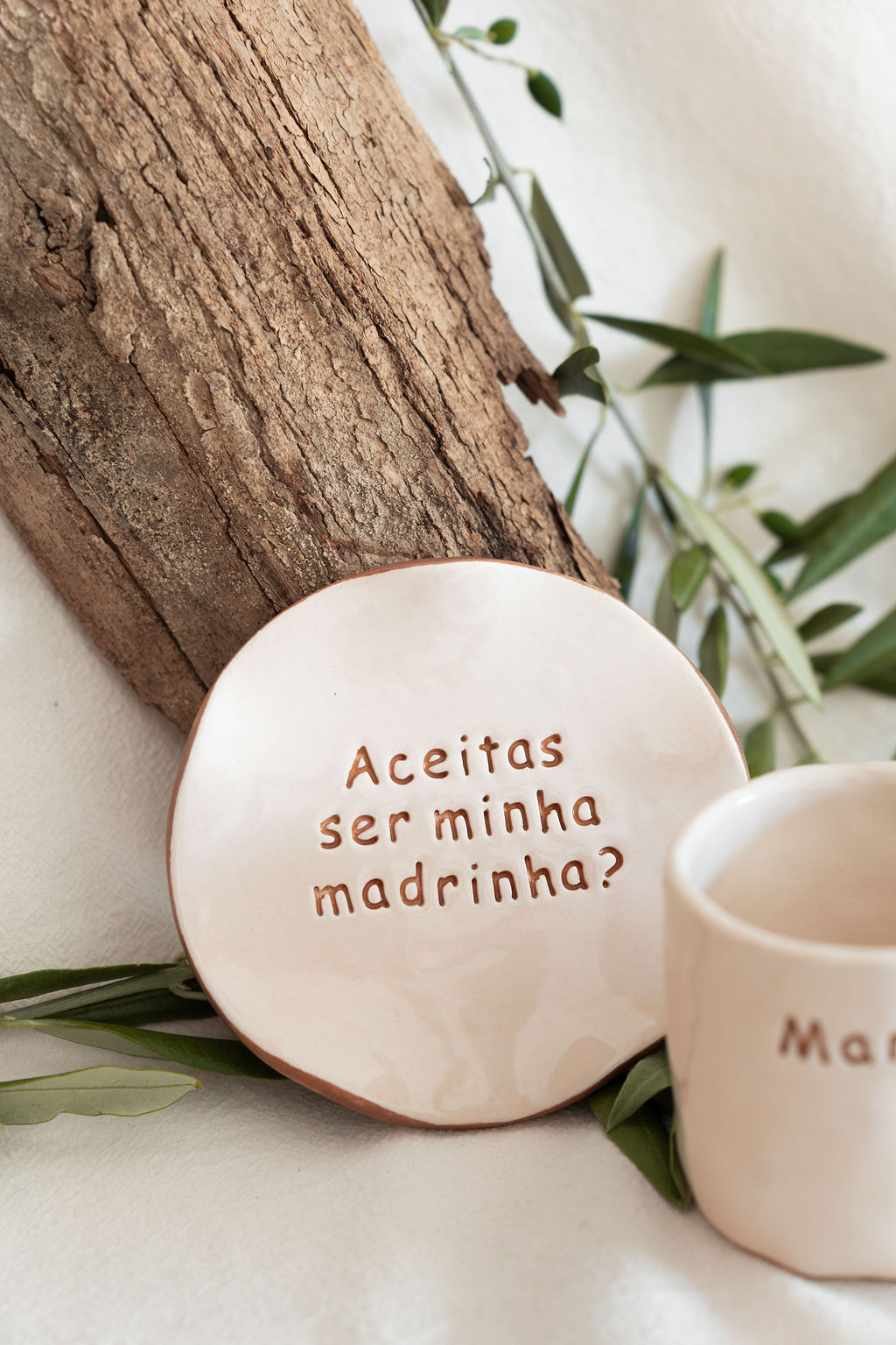 Prato redondo de cerâmica bege com texto Aceitas ser minha madrinha? encostado a casca de árvore