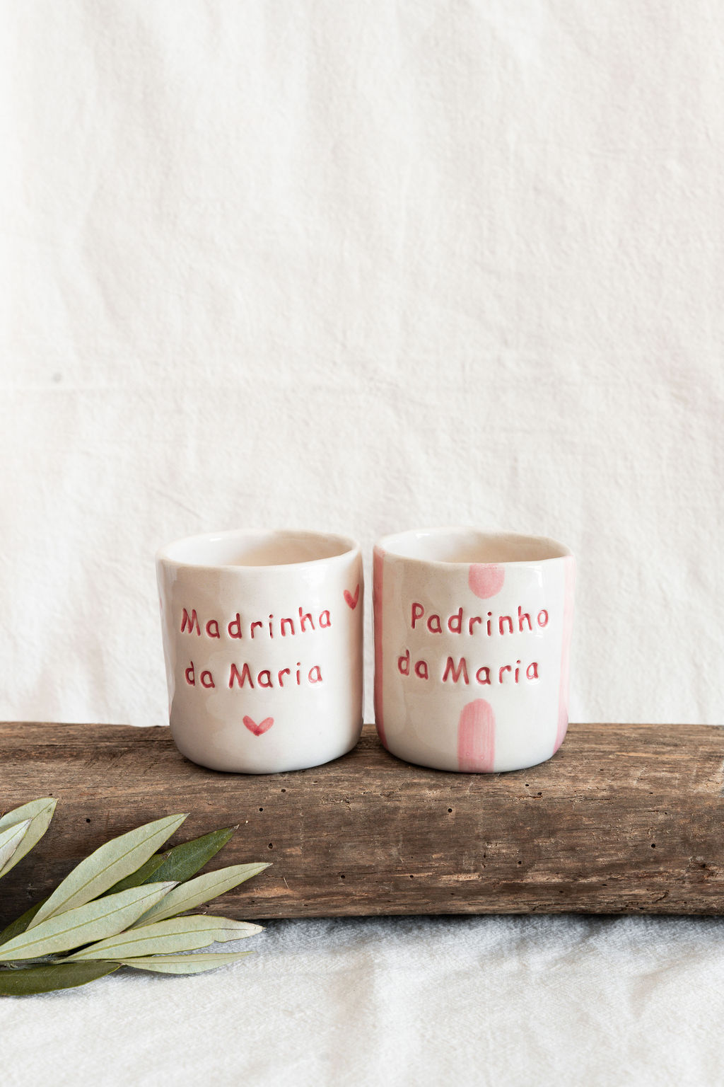 Dois copos de cerâmica artesanal com inscrições em português sobre um pedaço de madeira e fundo branco.
