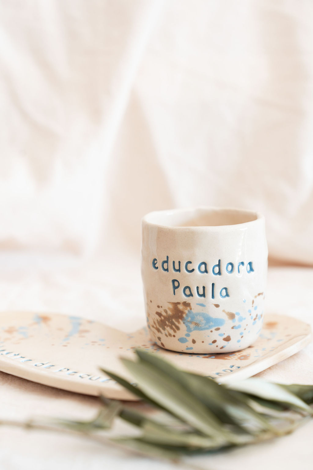 Caneca de cerâmica branca com texto azul educadora Paula e prato decorativo ao lado.