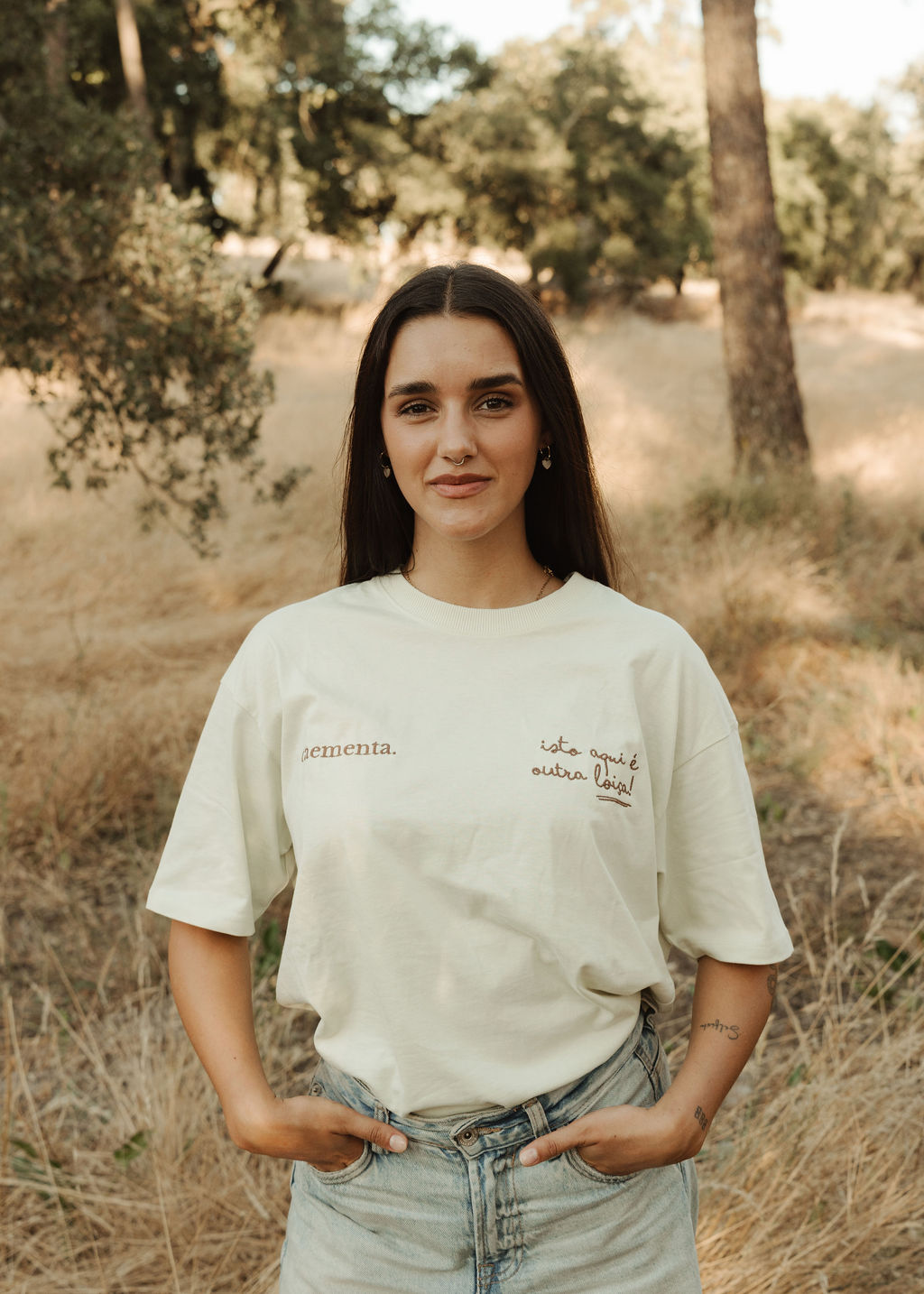 Mulher com t-shirt branca com texto bordado castanho e calças de ganga num ambiente campestre seco.