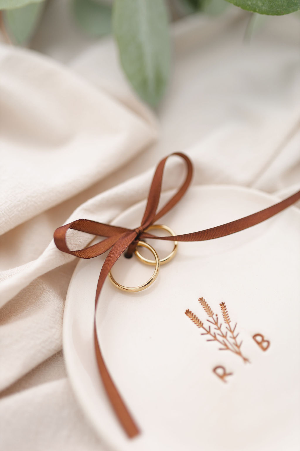 Prato branco com espigas e letras R B, alianças douradas com fita castanha e tecido bege.