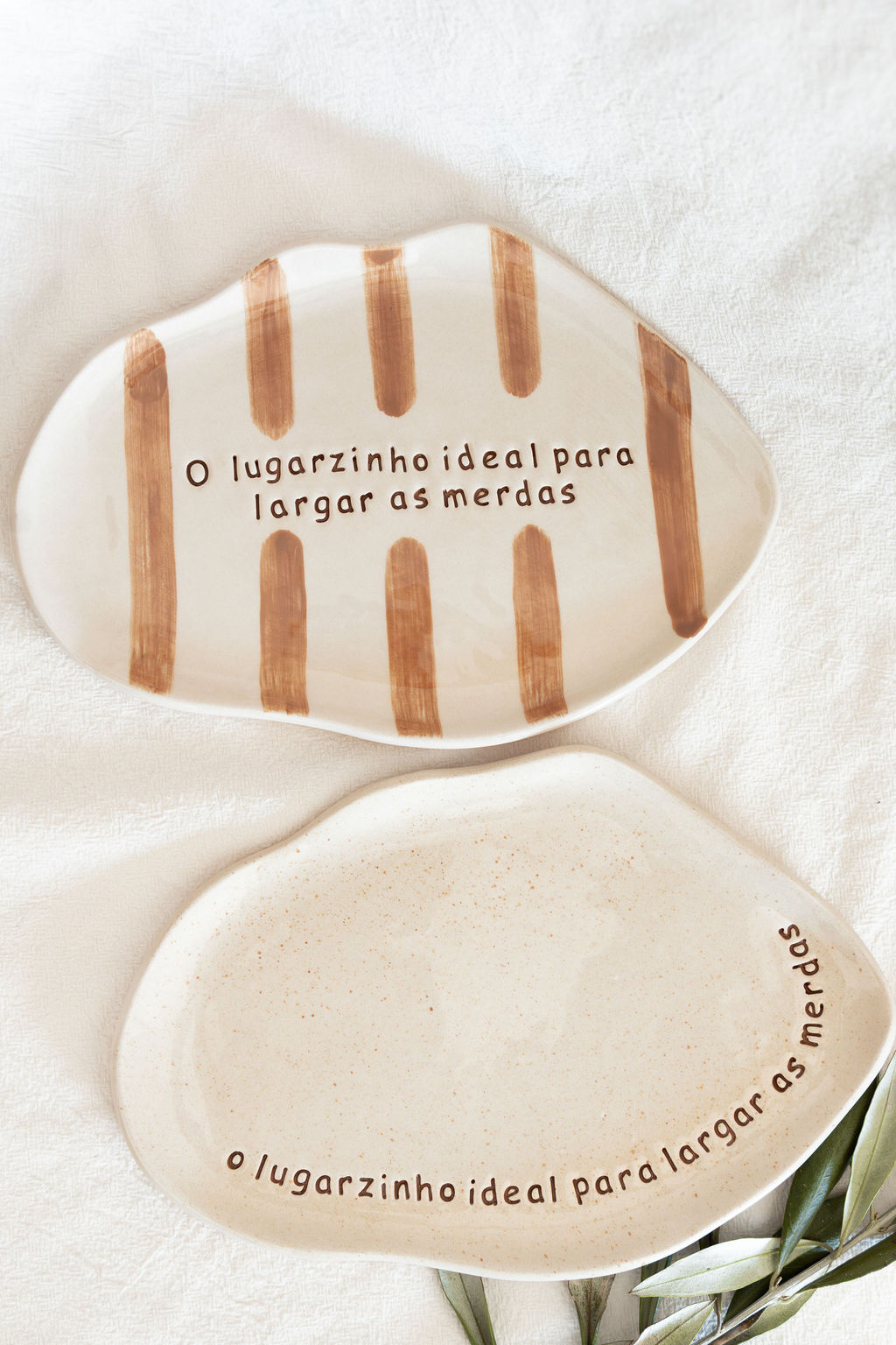 Duas travessas cerâmicas bege com texto em castanho sobre tecido branco e folhas verdes.