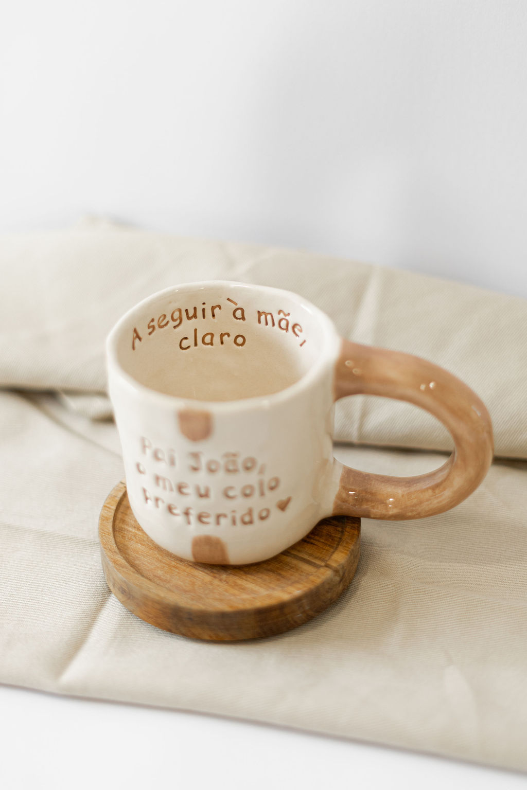 Caneca cerâmica creme com texto em castanho, pega castanha e base de madeira sobre tecido bege