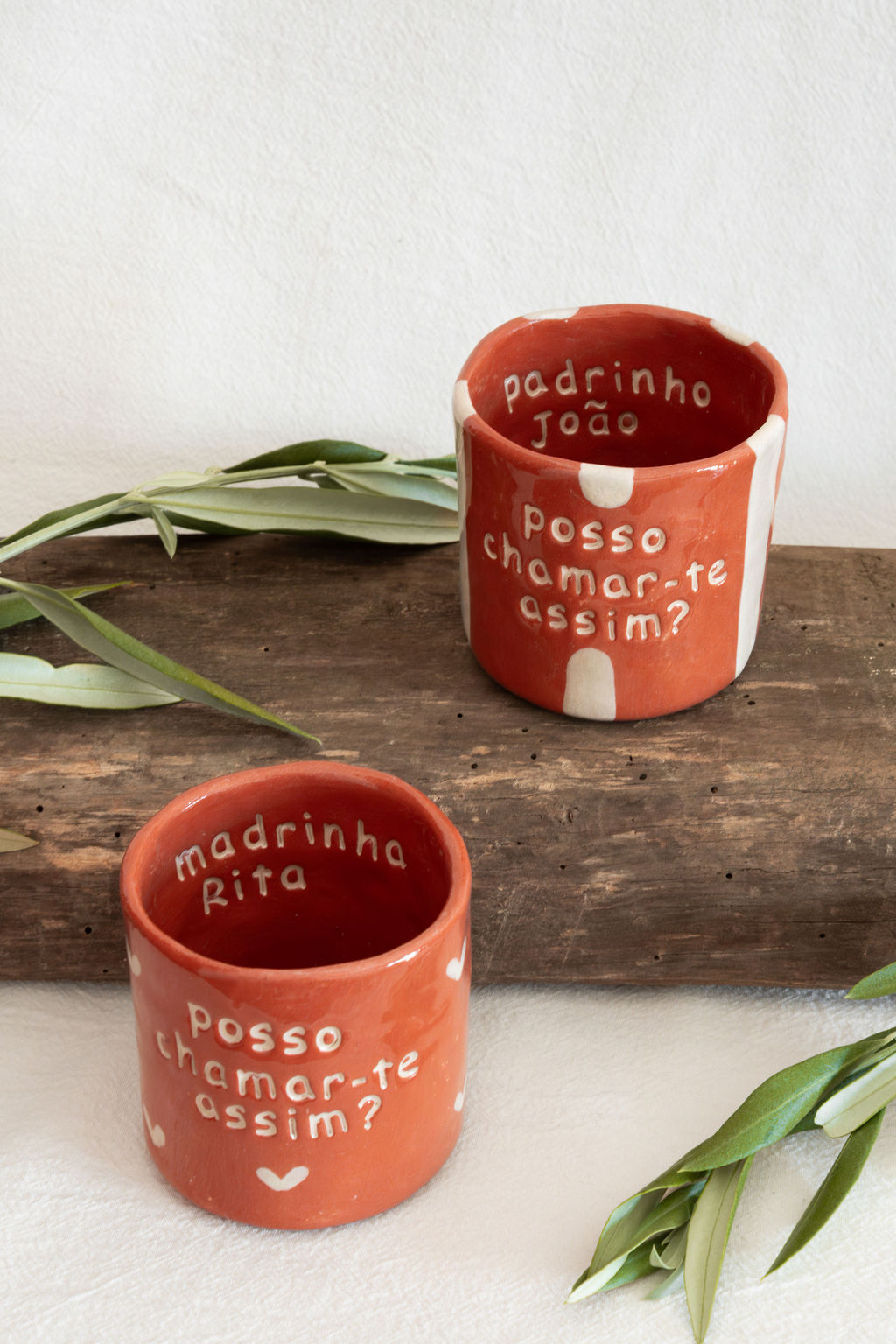 Dois vasos de cerâmica vermelhos com textos personalizados em português sobre uma tábua de madeira e folhas verdes no fundo branco.