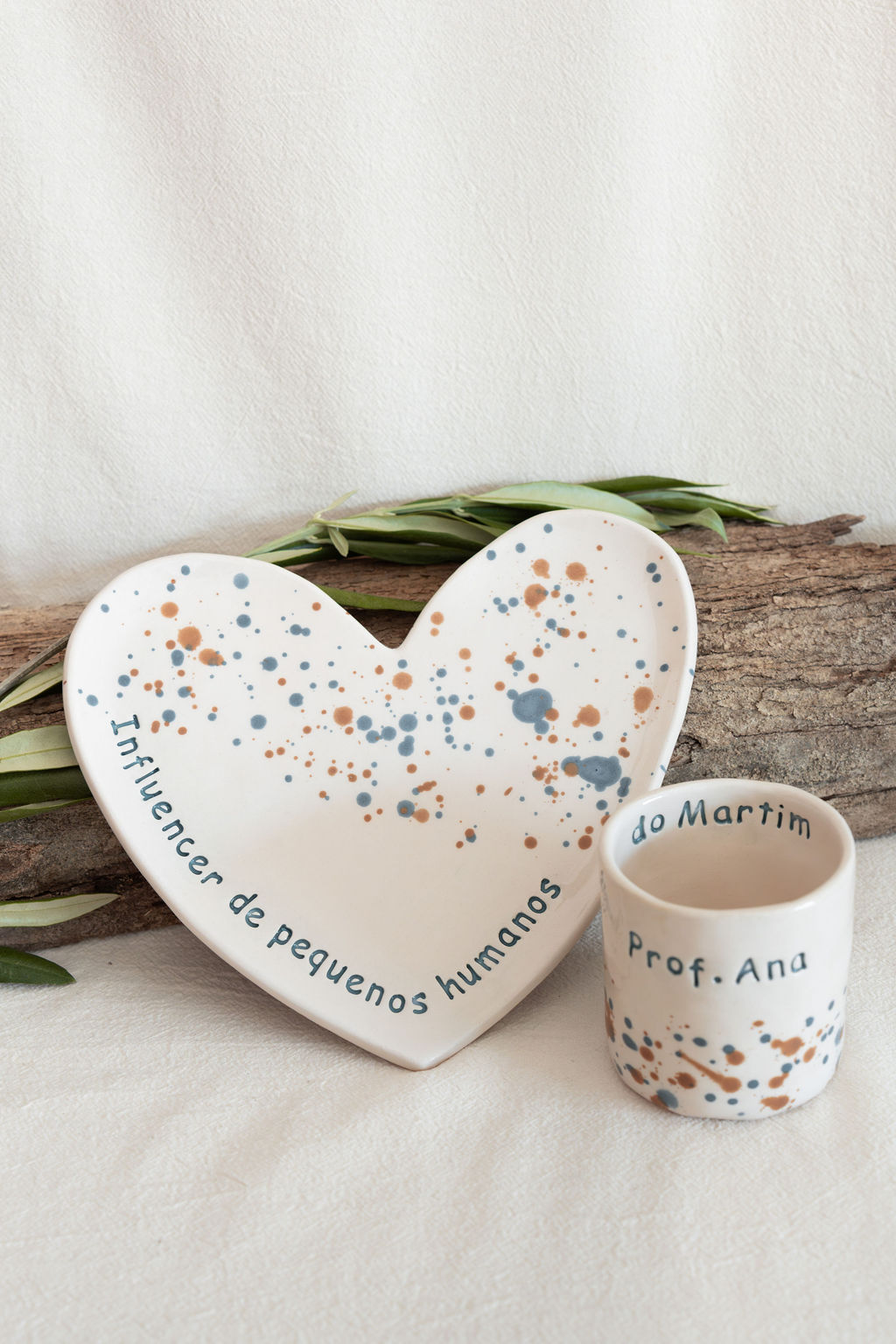 Conjunto cerâmico com prato coração e copo branco manchado com texto visível, decorado com tronco e folhas
