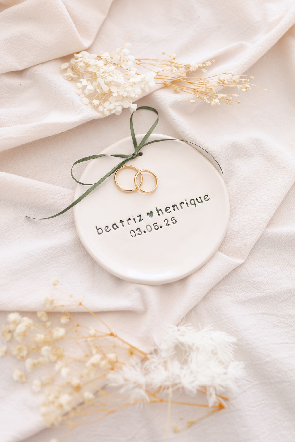 Prato cerâmico branco com duas alianças douradas e texto personalizado sobre pano branco e flores secas