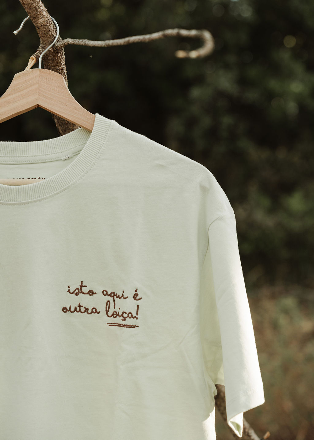 Camiseta bege clara com texto bordado pendurada num cabide numa árvore