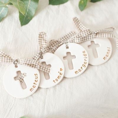 Ornamentos redondos de cerâmica branca com cruz e nomes, com laços xadrez castanhos e brancos, sobre tecido creme e folhas verdes