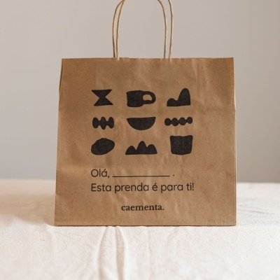 Saco de papel castanho com ilustrações pretas e texto em português colocado sobre tecido branco.