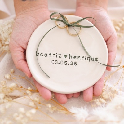 Prato cerâmico branco com nomes e data, alianças douradas com fita verde, mãos tatuadas seguram o prato