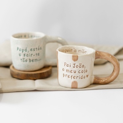 Duas canecas de cerâmica com frases em português colocadas sobre tecido bege e descanso de madeira.