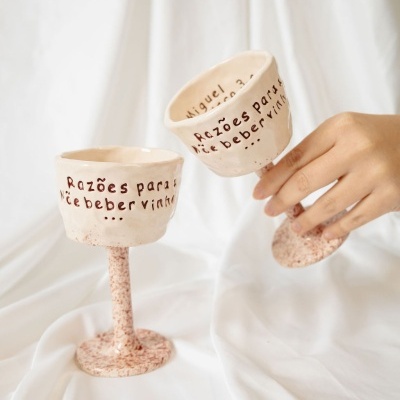 Duas taças de vinho em cerâmica com inscrição em português e acabamento craquelado, uma sendo segurada por uma mão