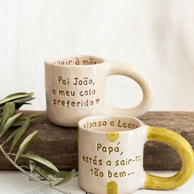 Duas canecas de cerâmica bege com texto em português, uma com alça amarela, em fundo bege e madeira com ramo verde